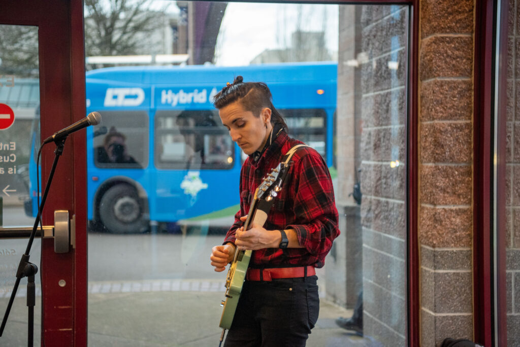 A musician in a red plaid shirt plays an electric guitar near a microphone stand, with a blue public transit bus visible through a window in the background.