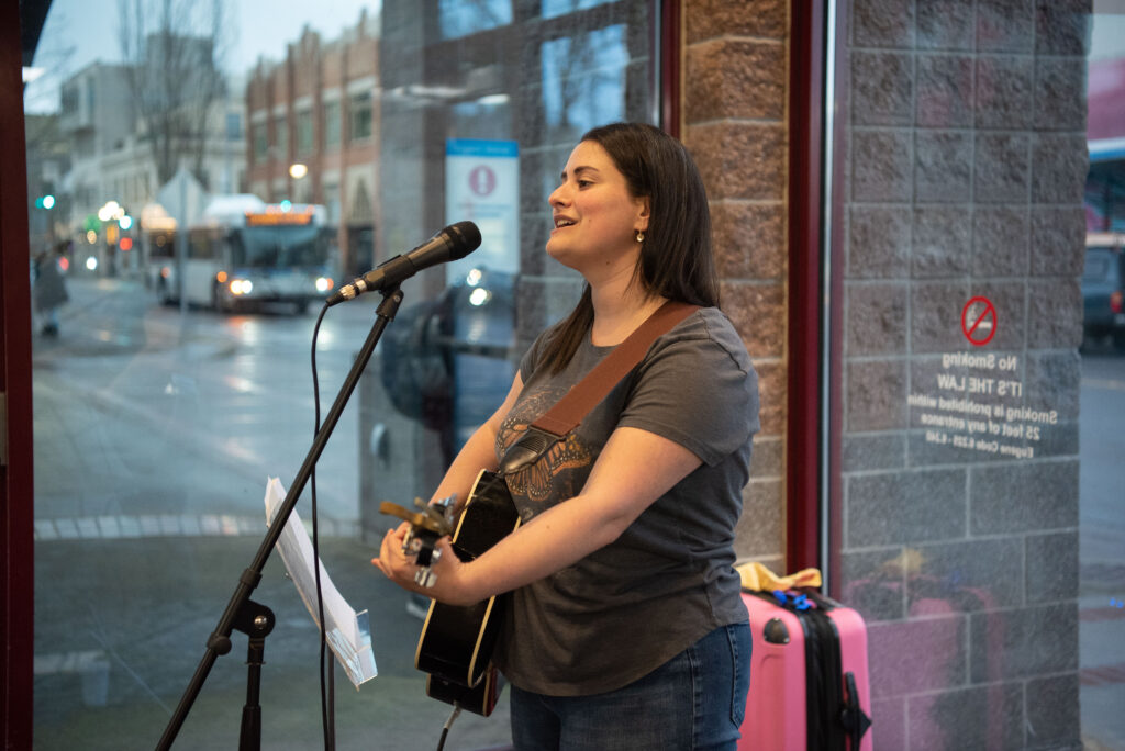 A woman playing an acoustic guitar and singing into a microphone in front of a window.