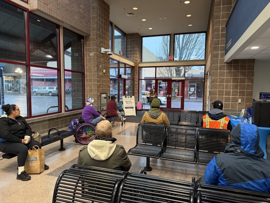 People sitting in a waiting area while a woman stands by the entrance holding a small sign.